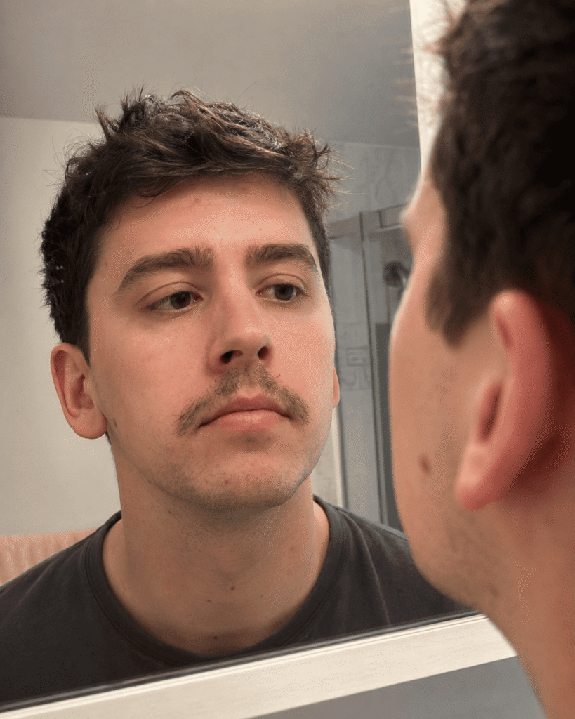 A realistic candid photo of a regular man in his late 20s with a 3-week-old patchy mustache, looking slightly unsure at himself in a mirror, natural indoor lighting, plain home bathroom background softly out of focus. Authentic skin, the mustache visibly uneven and a bit sparse in places — not perfect, not glamorous. Documentary-style, shot like a real smartphone photo, neutral expression, no retouching, true-to-life imperfections.